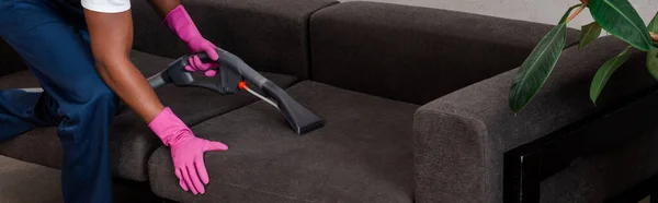 Panoramic shot of african american cleaner cleaning couch with vacuum cleaner at home — Stock Photo