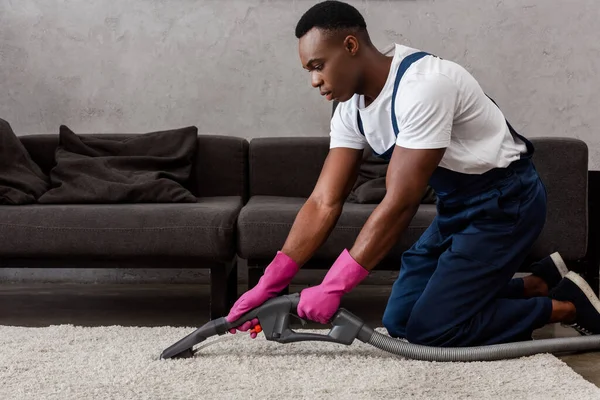 Nettoyant afro-américain dans des gants en caoutchouc nettoyage tapis dans le salon — Photo de stock