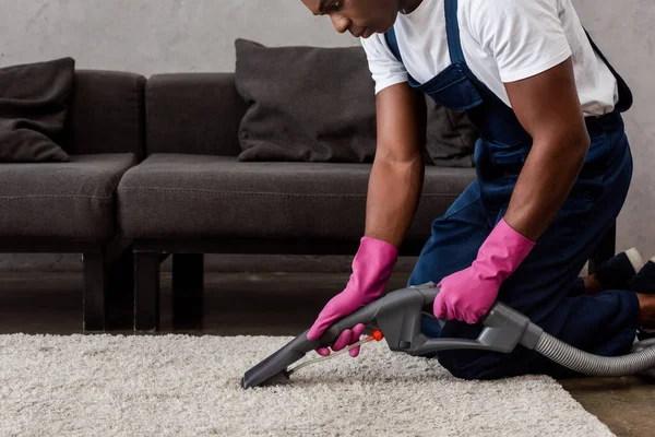 Vue recadrée du nettoyeur afro-américain utilisant un aspirateur sur tapis dans le salon — Photo de stock