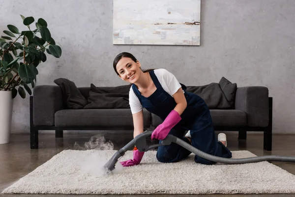 Limpiador sonriendo a la cámara mientras usa aspiradora con vapor caliente en la alfombra en la sala de estar - foto de stock