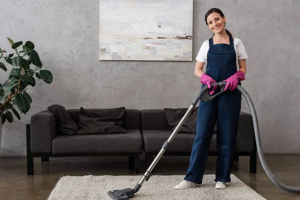 Sonriente limpiador en uniforme mirando a la cámara mientras se utiliza la aspiradora en casa - foto de stock