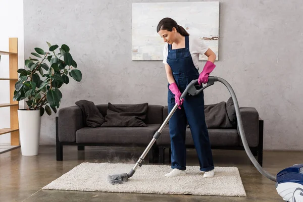 Hermosa limpiadora en ropa de trabajo y guantes de goma usando aspiradora con vapor caliente en la alfombra en la sala de estar - foto de stock