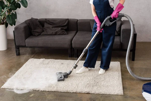 Vista recortada de la limpiadora en overoles y guantes de goma usando aspiradora con vapor caliente en la alfombra — Stock Photo