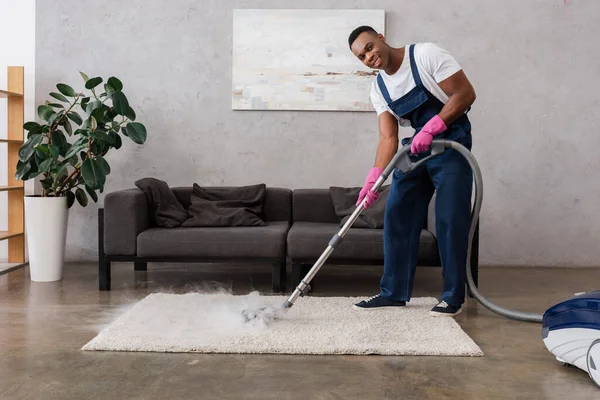 Sonriente limpiador afroamericano en overoles usando aspiradora con vapor caliente en casa - foto de stock