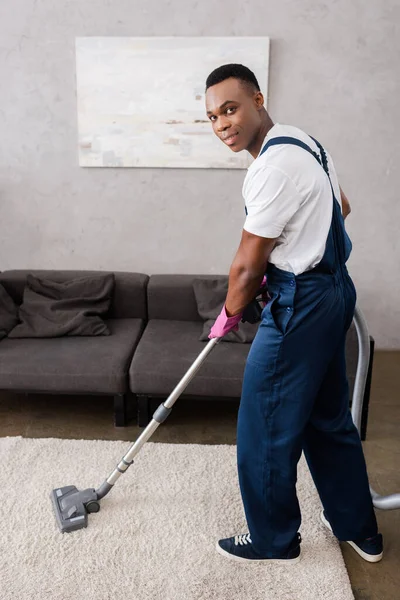 African american cleaner in uniform cleaning carpet and looking at camera in living room — Stock Photo