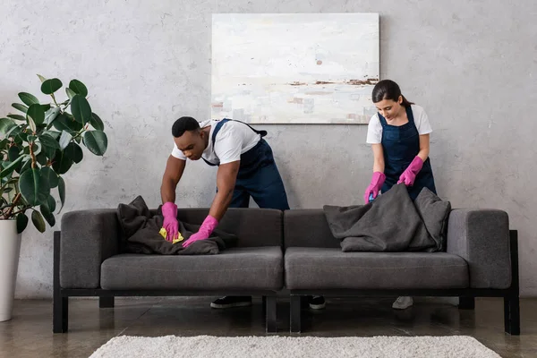 Nettoyeurs multiculturels dans le canapé de nettoyage uniforme avec chiffons dans le salon — Photo de stock