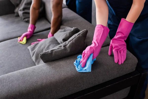 Cropped view of multicultural cleaners in rubber gloves cleaning couch — Stock Photo