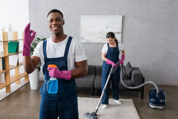 Selective focus of smiling african american cleaner showing okay and holding detergent near colleague using vacuum cleaner — Stock Photo