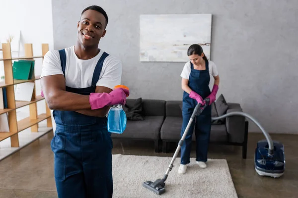 Focus selettivo del detergente africano americano con detergente e sorridente davanti alla telecamera vicino al tappeto di pulizia del collega — Foto stock