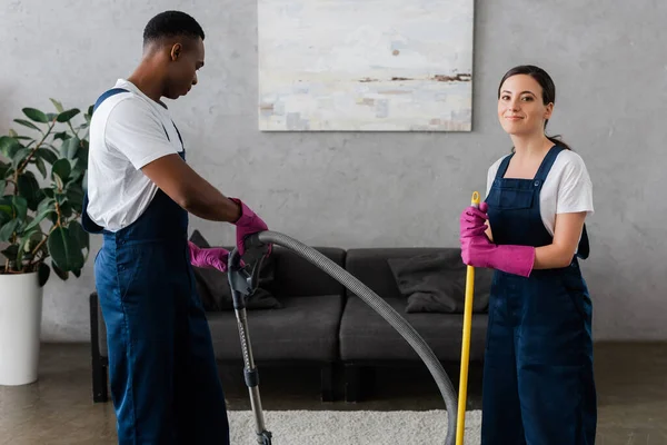 Lächelnde Putzfrau in Uniform blickt in die Kamera neben einem amerikanischen Kollegen mit Staubsauger — Stockfoto