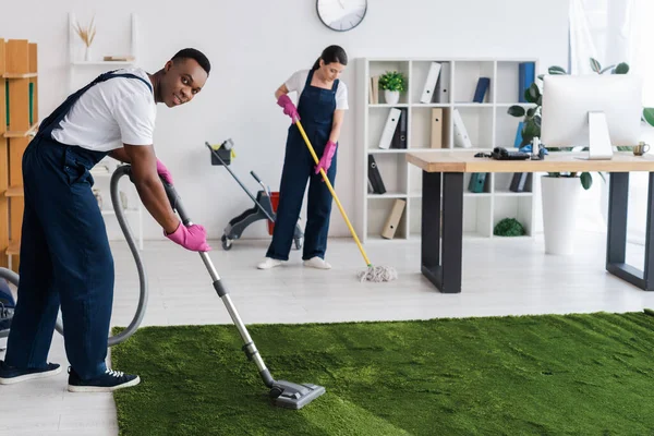 Selective focus of smiling african american cleaner vacuuming carpet near colleague with mop in office — Stock Photo