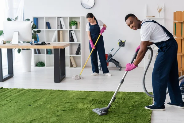 Selektiver Fokus einer afrikanisch-amerikanischen Putzfrau, die lächelt, während sie Teppich in der Nähe ihres Kollegen mit einem Wischmopp putzt — Stockfoto