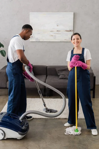 Limpiador sonriente sosteniendo fregona cerca de un colega afroamericano limpiando alfombras en casa - foto de stock