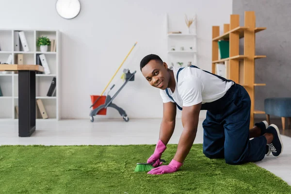 Sonriente limpiador afroamericano sosteniendo cepillo mientras limpia la alfombra en la oficina - foto de stock