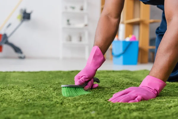 Vista recortada de limpiador afroamericano en guantes de goma alfombra de limpieza con cepillo en la oficina - foto de stock