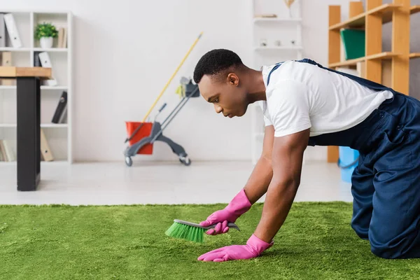 Vista lateral de limpiador afroamericano en uniforme y guantes de goma limpiando alfombra con cepillo en oficina - foto de stock