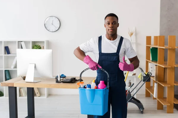 Nettoyant afro-américain montrant pouce levé et tenant seau de fournitures de nettoyage dans le bureau — Photo de stock