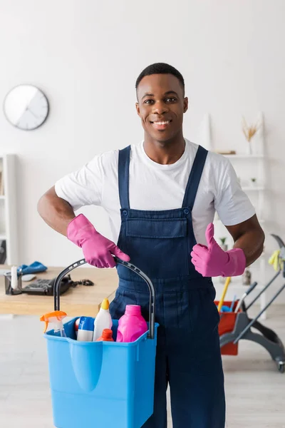 Lächelnde afrikanisch-amerikanische Putzfrau in Uniform zeigt wie und hält Eimer mit Putzmitteln im Büro — Stockfoto