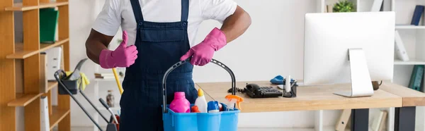 Cultura panorâmica de limpador afro-americano em uniforme mostrando como e segurando suprimentos de limpeza em balde no escritório — Fotografia de Stock