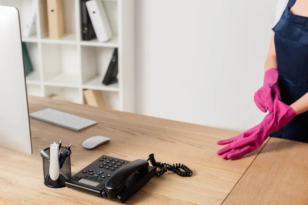 Ausgeschnittene Ansicht einer Reinigungskraft mit Gummihandschuhen in der Nähe von Computer und Telefon auf dem Bürotisch — Stockfoto