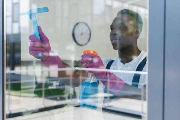 Selective focus of african american cleaner in rubber gloves using detergent and squeegee handle while cleaning window — Stock Photo