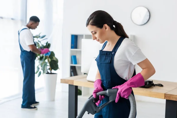Selective focus of attractive worker of cleaning service using vacuum cleaner near african american colleague in office — Stock Photo