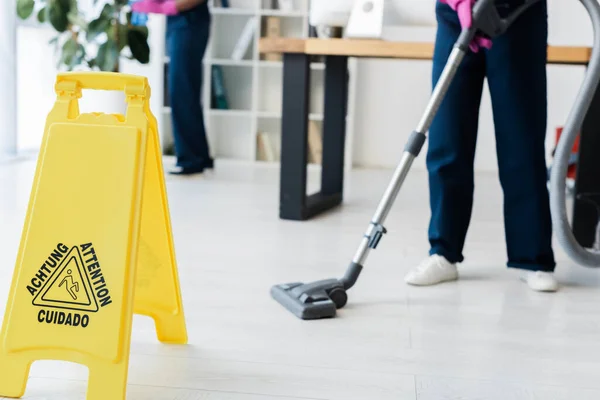 Cropped view of cleaners working in office near wet floor sign with attention lettering on floor — Stock Photo