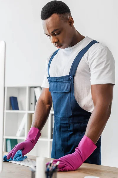 Selektiver Fokus der afrikanisch-amerikanischen Reinigungskraft in Overalls, die Computertastatur im Büro reinigt — Stockfoto