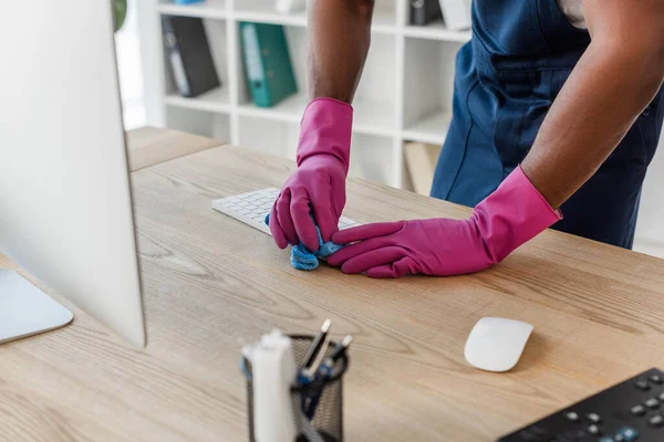 Selektiver Fokus der afrikanisch-amerikanischen Reinigungskraft mit Lappen beim Reinigen der Computertastatur auf dem Tisch im Büro — Stockfoto