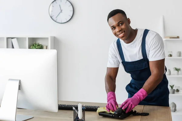 Selective focus of smiling african american cleaner cleaning telephone with rag on office table — Stock Photo