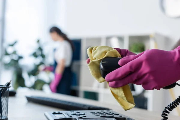 Selective focus of cleaner in rubber gloves cleaning telephone near colleague in office — Stock Photo