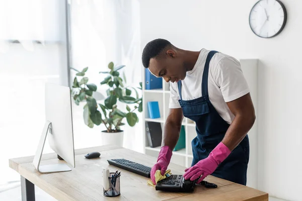 Afroamerikanische Reinigungskraft benutzt Lappen beim Putzen des Telefons in der Nähe des Computers auf dem Bürotisch — Stockfoto