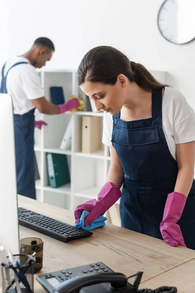 Selektiver Fokus der Reinigungskraft mit Lappen im Büro in der Nähe eines afrikanischen amerikanischen Kollegen — Stockfoto