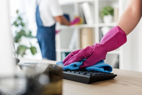 Vista recortada del limpiador frotando el teclado de la computadora en la mesa cerca de un colega afroamericano en la oficina - foto de stock