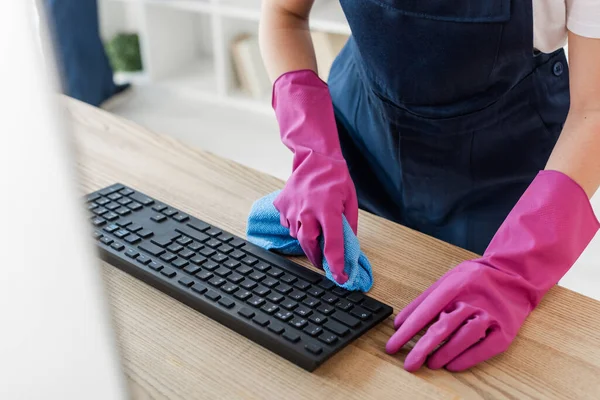 Vista cortada de limpador em luvas de borracha segurando pano perto do teclado do computador no escritório — Fotografia de Stock