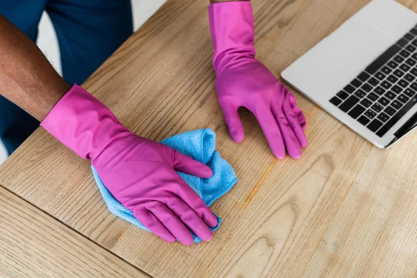 Vue recadrée de nettoyant afro-américain dans des gants en caoutchouc table de nettoyage près d'un ordinateur portable dans le bureau — Photo de stock