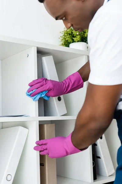 Selective focus of african american cleaner in rubber gloves cleaning cupboard near paper folders in office — Stock Photo