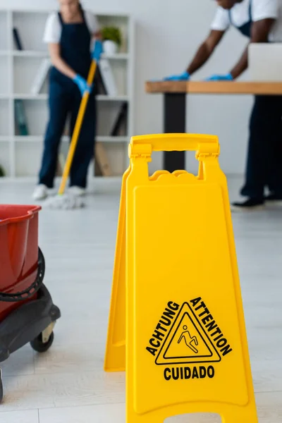 Focus selettivo del segno pavimento bagnato con lettere di attenzione e pulitori multietnici che lavorano in ufficio — Foto stock