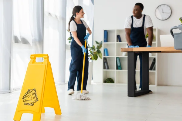 Selective focus of wet floor sign and smiling multiethnic cleaners working in office — Stock Photo