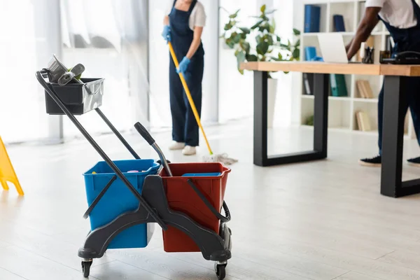 Enfoque selectivo de cubos con artículos de limpieza y limpiadores multiétnicos que trabajan en la oficina - foto de stock