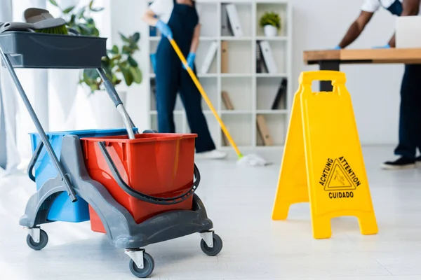 Selective focus of cleaning trolley near wet floor sign and multiethnic cleaners working in office — Stock Photo