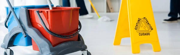 Récolte panoramique de chariot de nettoyage près des panneaux de sol mouillé et des nettoyeurs travaillant dans le bureau — Photo de stock