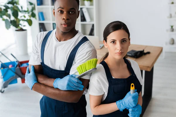 Multiethnic cleaners holding brush and mop while looking at camera in office — Stock Photo