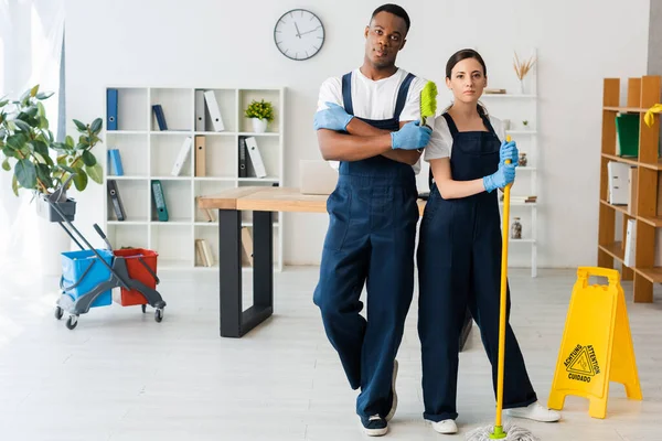 Pulitori multiculturali con scopa e spazzola guardando la fotocamera vicino al segno del pavimento bagnato in ufficio — Foto stock