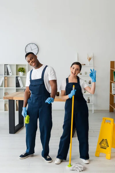 Multiethnic cleaners with brush and mop smiling at camera and showing ok gesture near wet floor sign in office — Stock Photo
