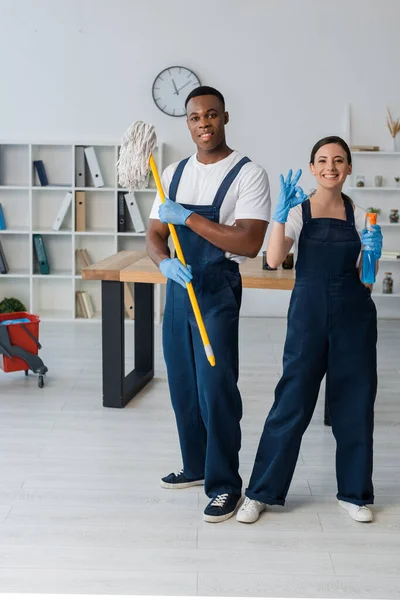 Sorrindo limpadores multiétnicos segurando esfregona e detergente e mostrando gesto ok no escritório — Fotografia de Stock