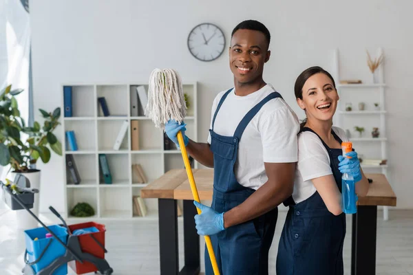 Positive multiethnic cleaners holding mop and detergent while smiling at camera in office — Stock Photo
