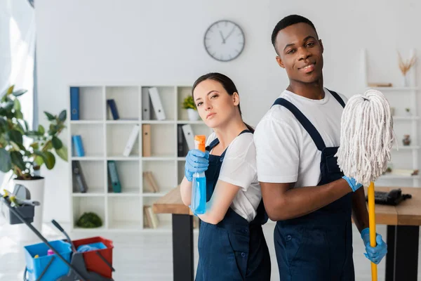 Nettoyant afro-américain souriant tenant une serpillière près d'un collègue avec du détergent au bureau — Photo de stock