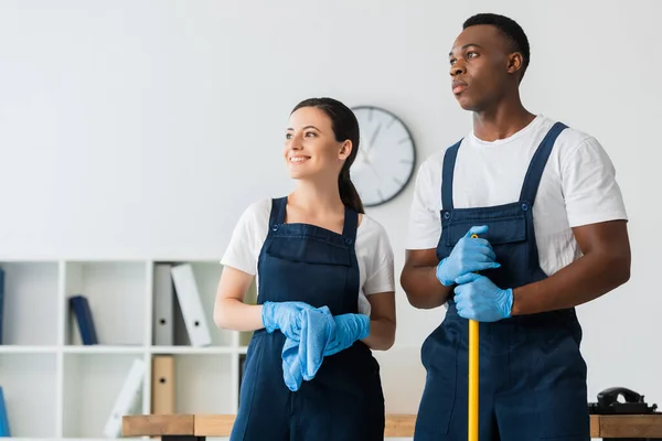 Sorrindo limpador segurando pano perto de colega afro-americano com esfregona no escritório — Fotografia de Stock