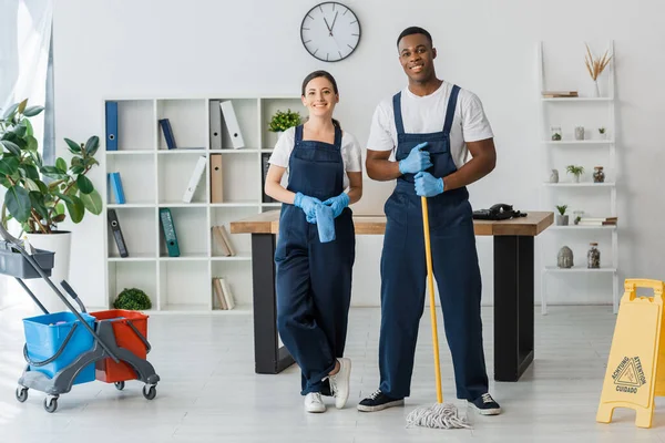 Multicultural cleaners with rag and mop smiling at camera near wet floor sign and cleaning trolley in office — Stock Photo